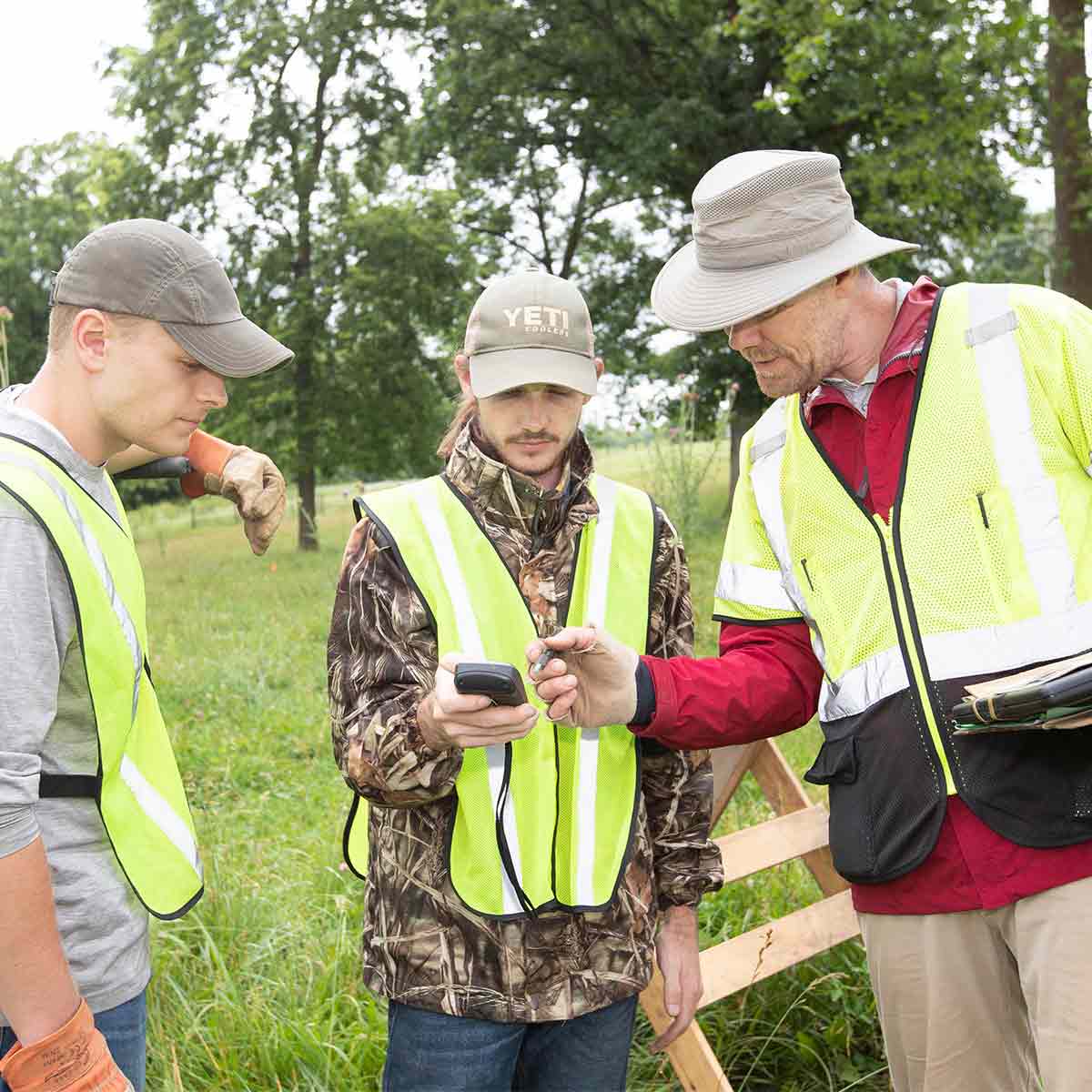 Two students and a professor wearing yellow safety vests have a discussion at an environmental site near Springfield.